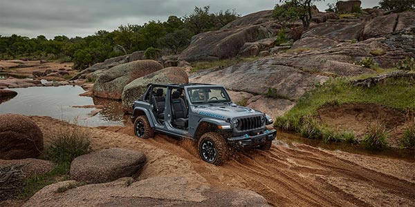 Profile view of a grey 2024 Jeep Wrangler 4xe driving on a dirt road with rocks and trees shown in the background.
