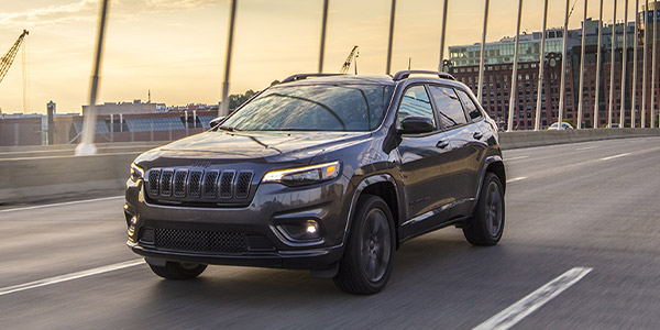 Front view of a dark grey 2023 Jeep Cherokee beind driven  on a suspension bridge with commercial buildings shown behind.