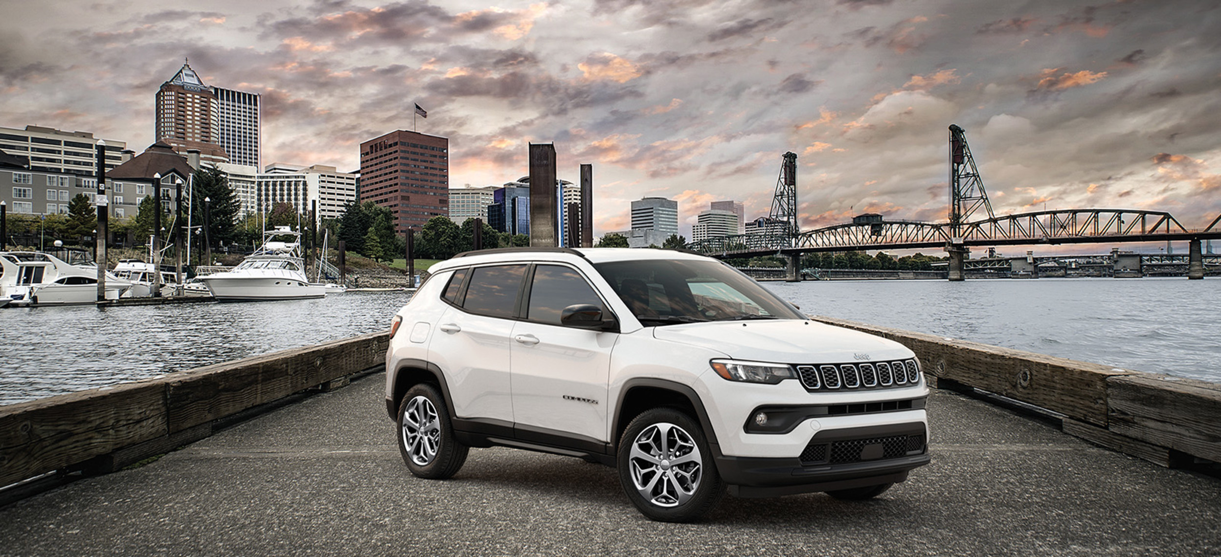 A profile view of a white 2024 Jeep Compass shown parked on a bridge suspended above water with a cityscape background. 