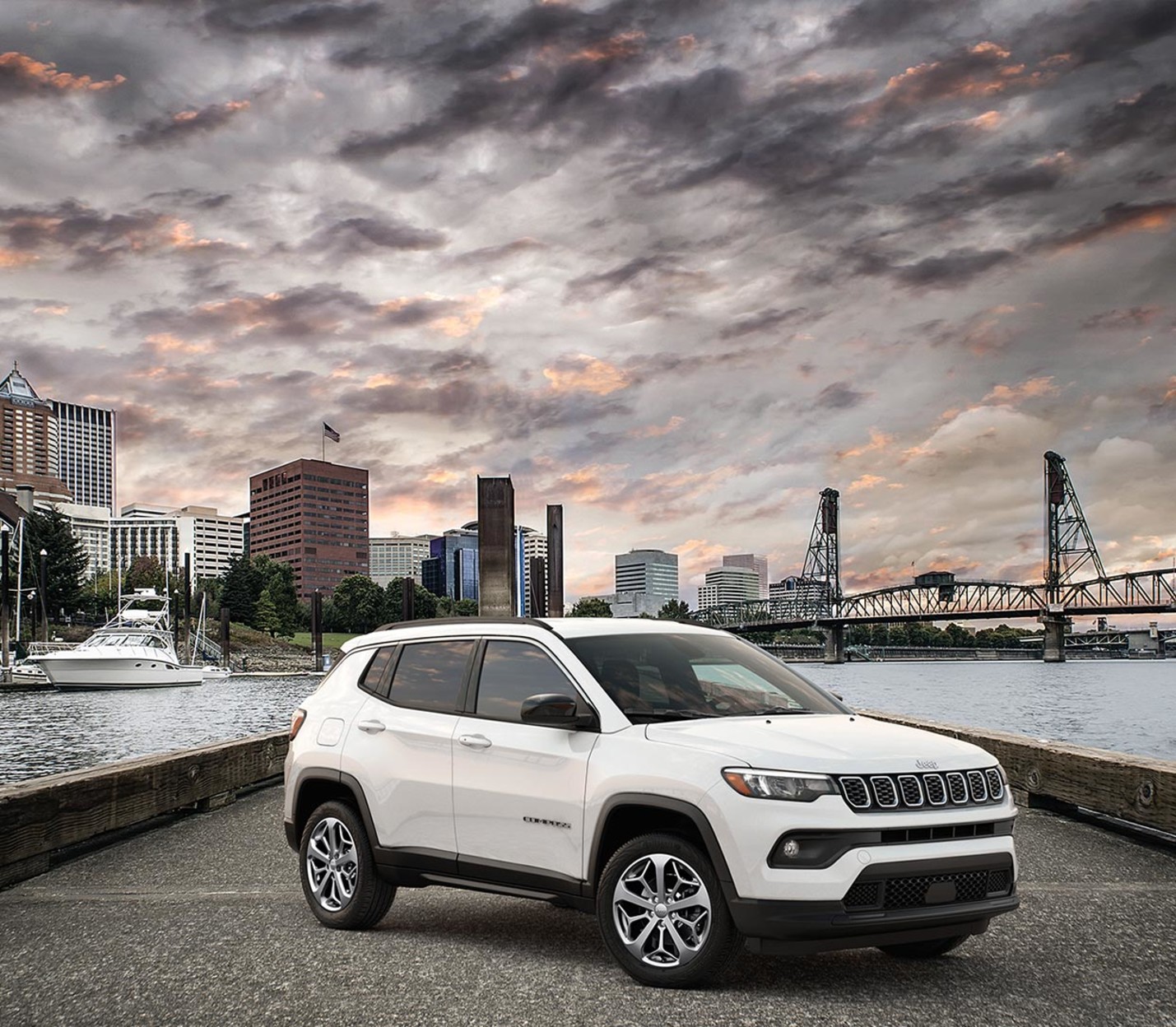 A profile view of a white 2024 Jeep Compass shown parked on a bridge suspended above water with a cityscape background. 