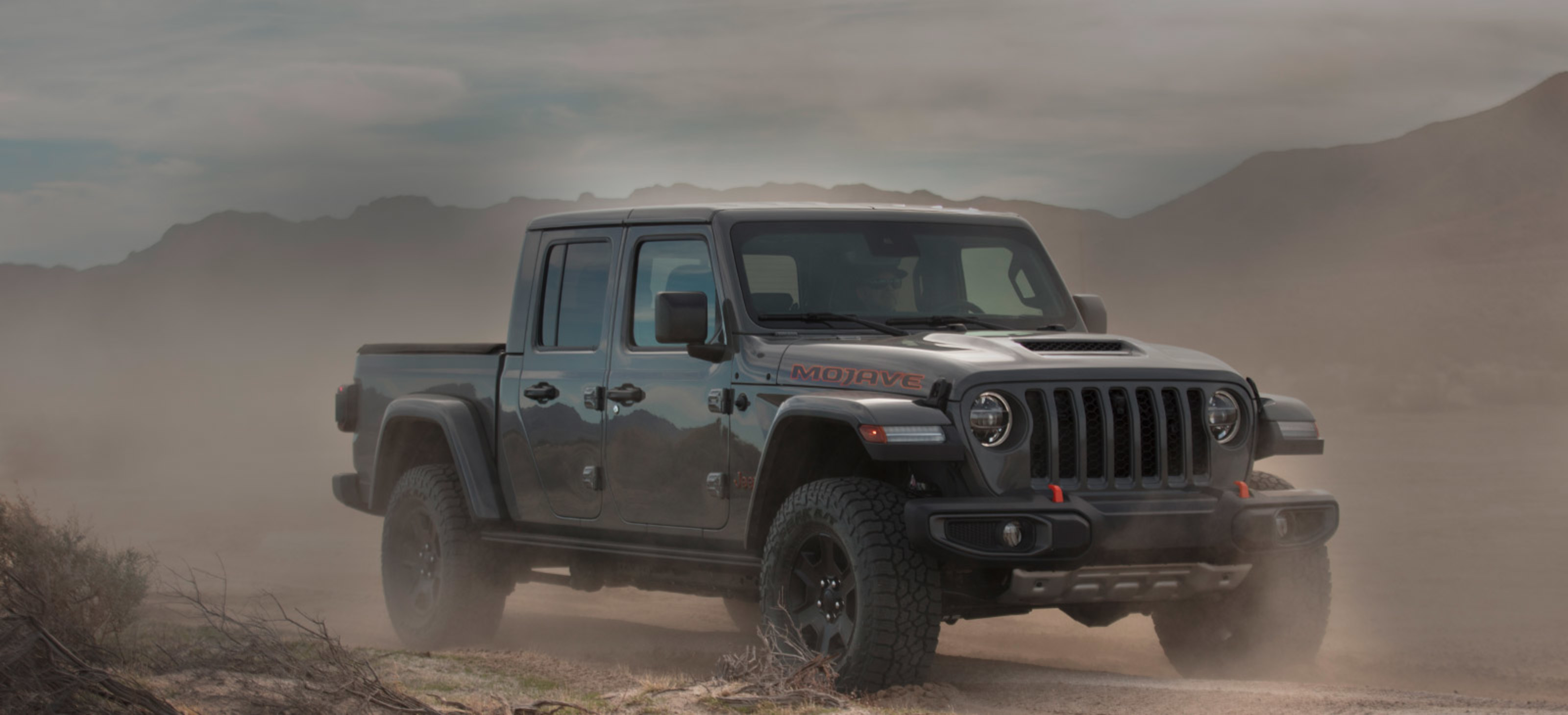 Vue avant d’un Jeep Gladiator Mojave 2021 gris pastenague roulant sur une route dans le désert, de la poussière tourbillonnant tout autour.