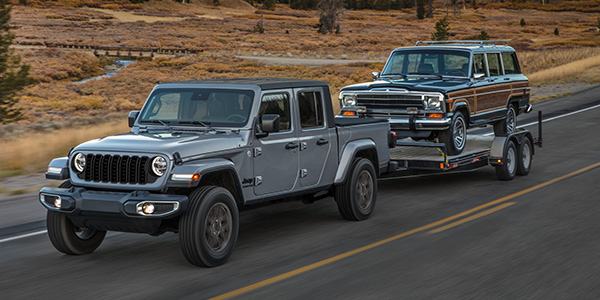 A 2024 Jeep Gladiator driving on a paved road in a desert landscape towing a vintage car on a flatbed trailer. 