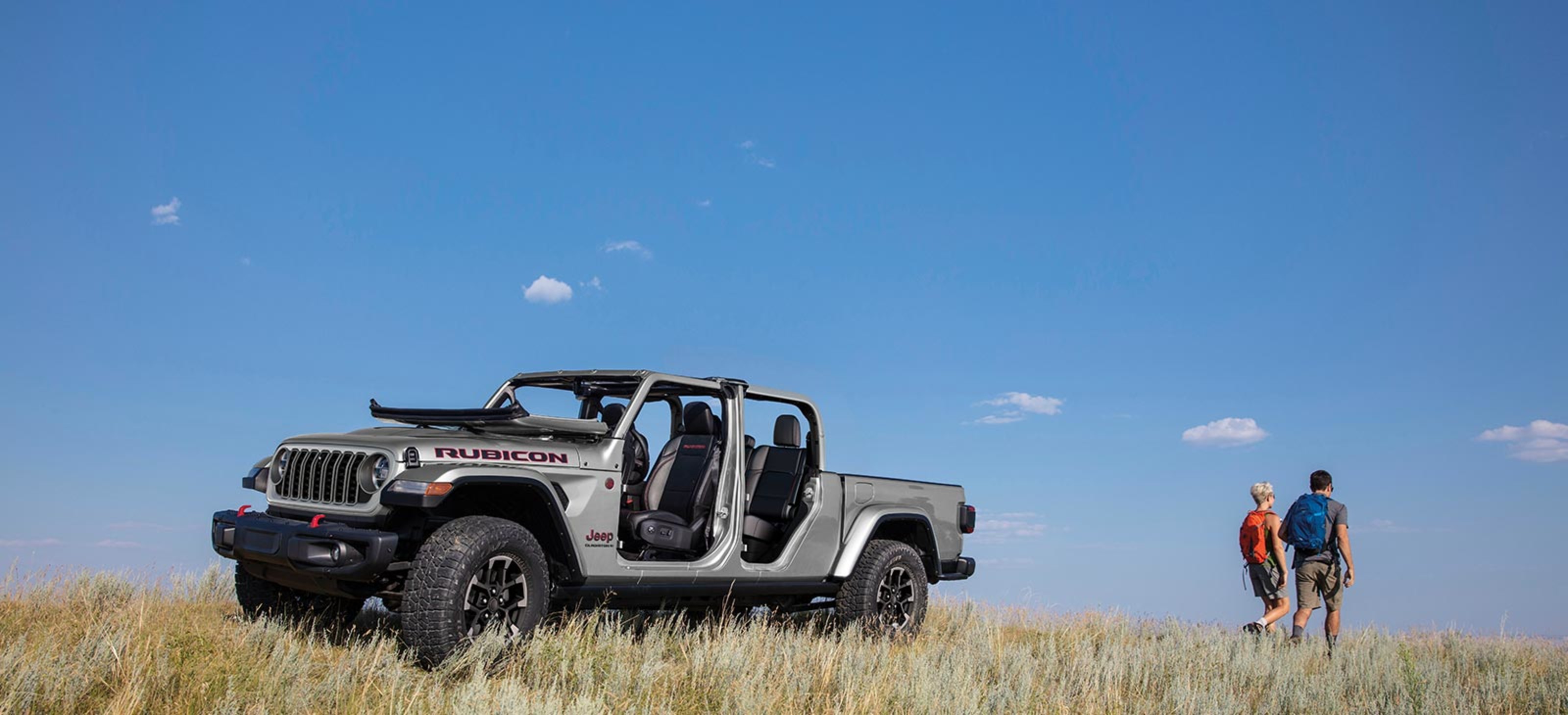 View of a silver 2024 Jeep Gladiator shown parked outside in tall grass and people walking away from the vehicle.