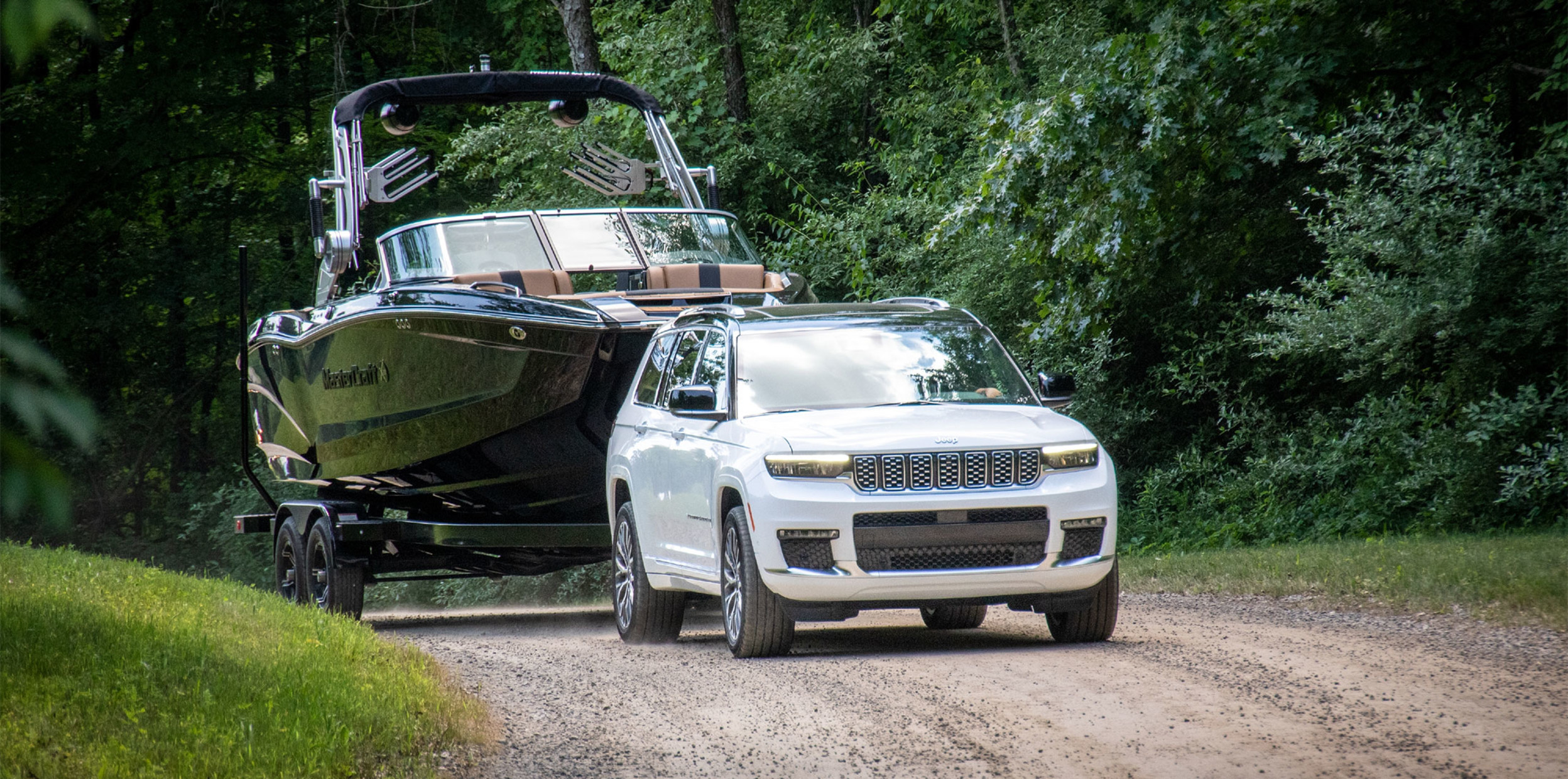 View of a white 2023 Jeep Grand Cherokee parked uphill on an unpaved road with a boat hitched to the back of the vehicle.