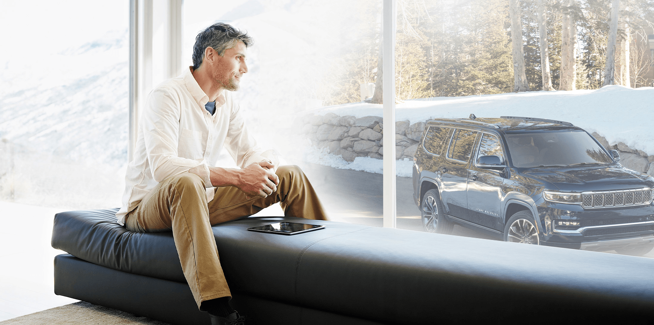A person sitting on a black couch indoors, looking out the window. A black All-New 2022 Grand Wagoneer is parked outside.