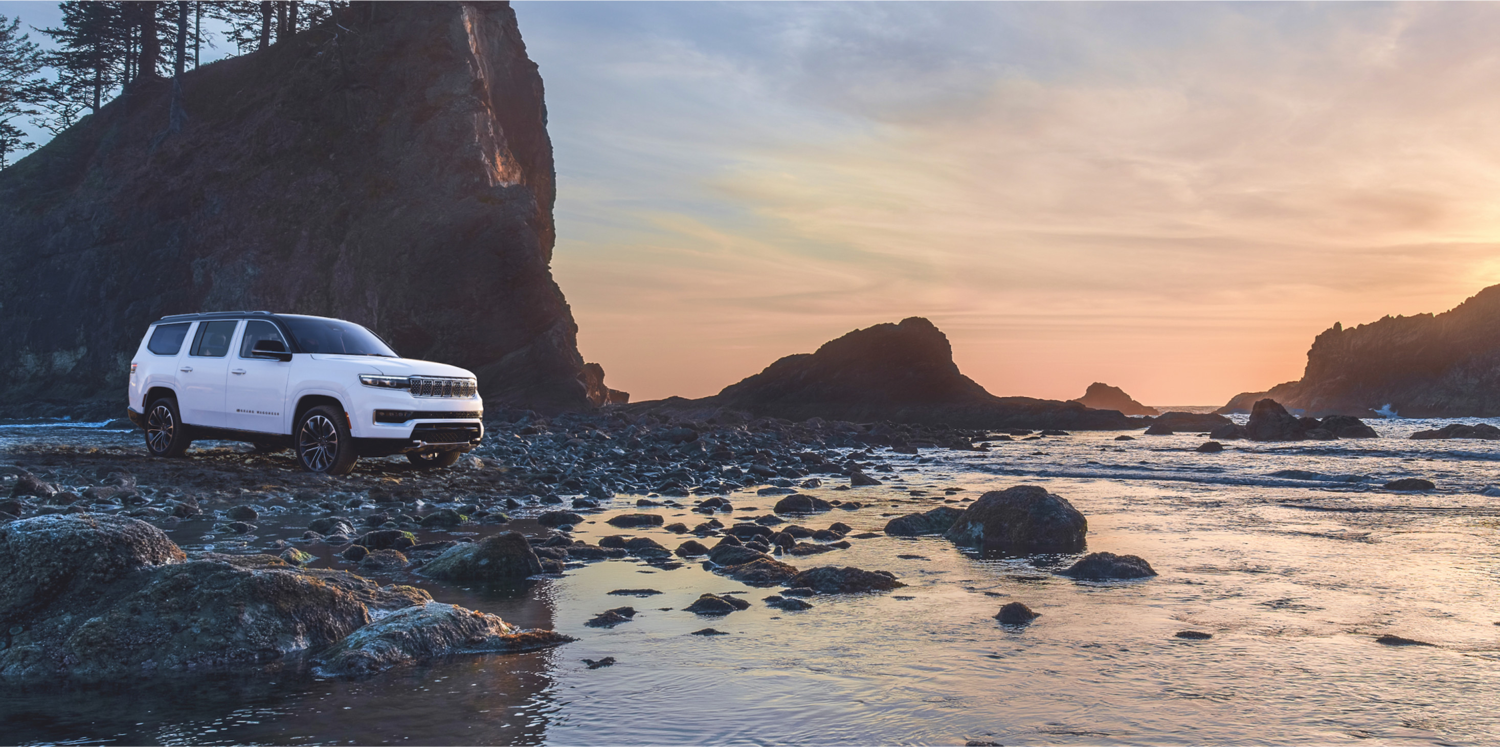 Jeep Grand Wagoneer 2024 blanc stationné sur un rivage rocheux au crépuscule.
