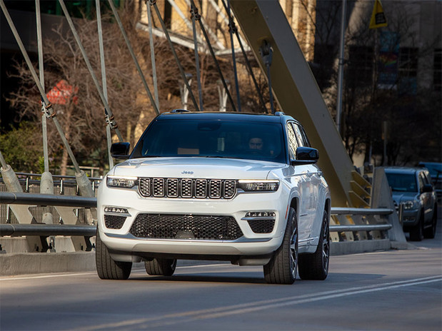 Front view of a white 2023 Jeep Grand Cherokee shown driving on a suspension bridge with buildings, trees and cars shown behind.