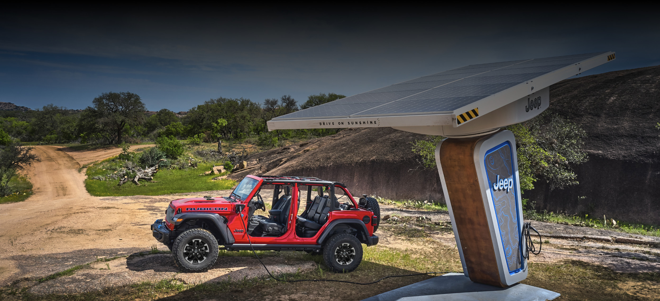 Red 2025 Jeep Wrangler Rubicon without a roof, parked near a solar-powered charging station in a natural outdoor setting.