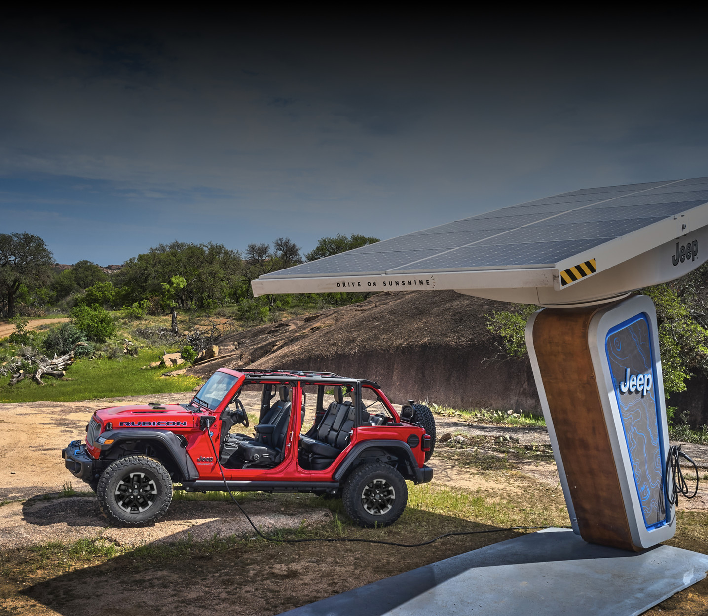 Red 2025 Jeep Wrangler Rubicon without a roof, parked near a solar-powered charging station in a natural outdoor setting.