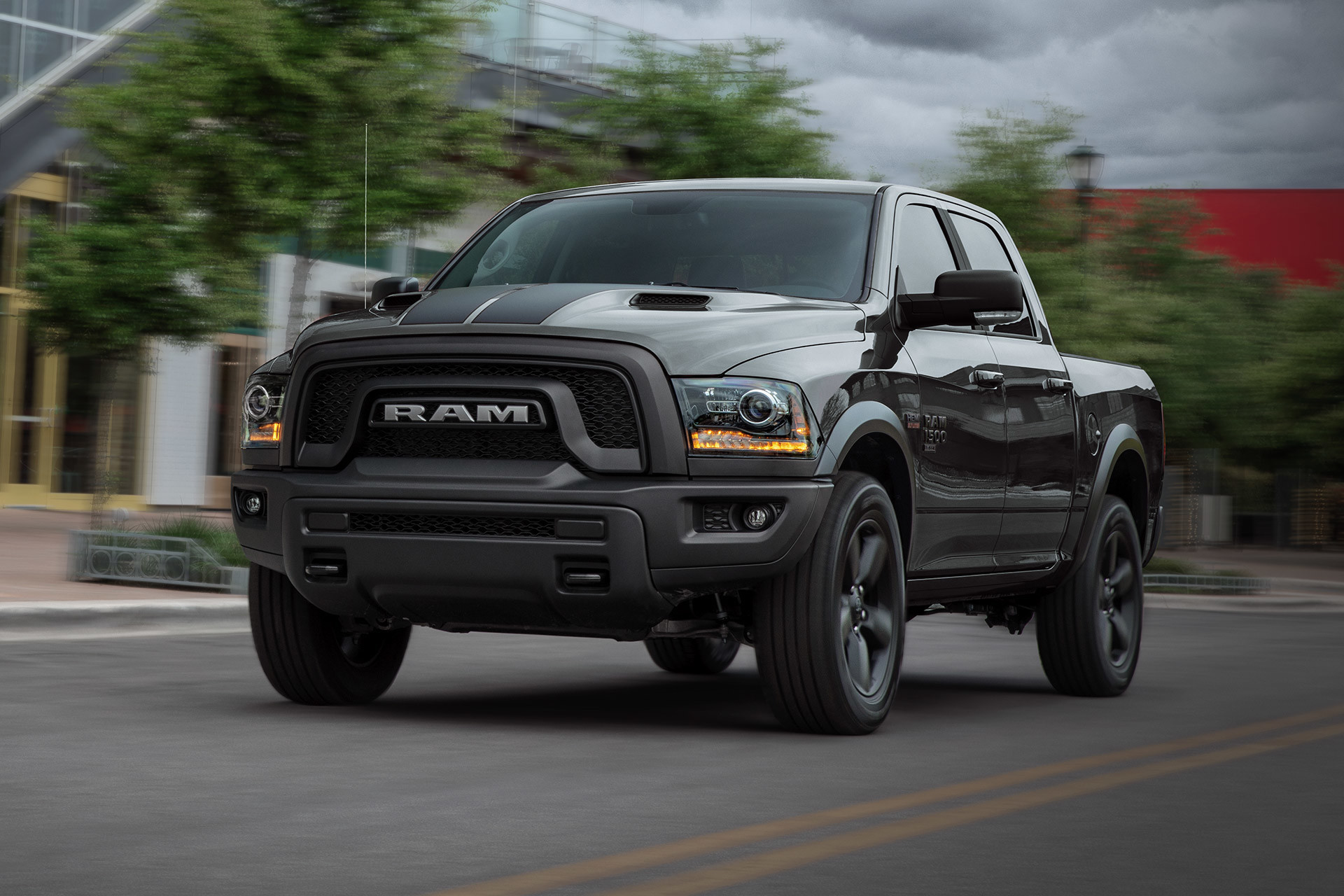  A front view of a Diamond Black Crystal Pearl 2021 Ram 1500 Classic Warlock being driven on a road in front of a glass-walled building.