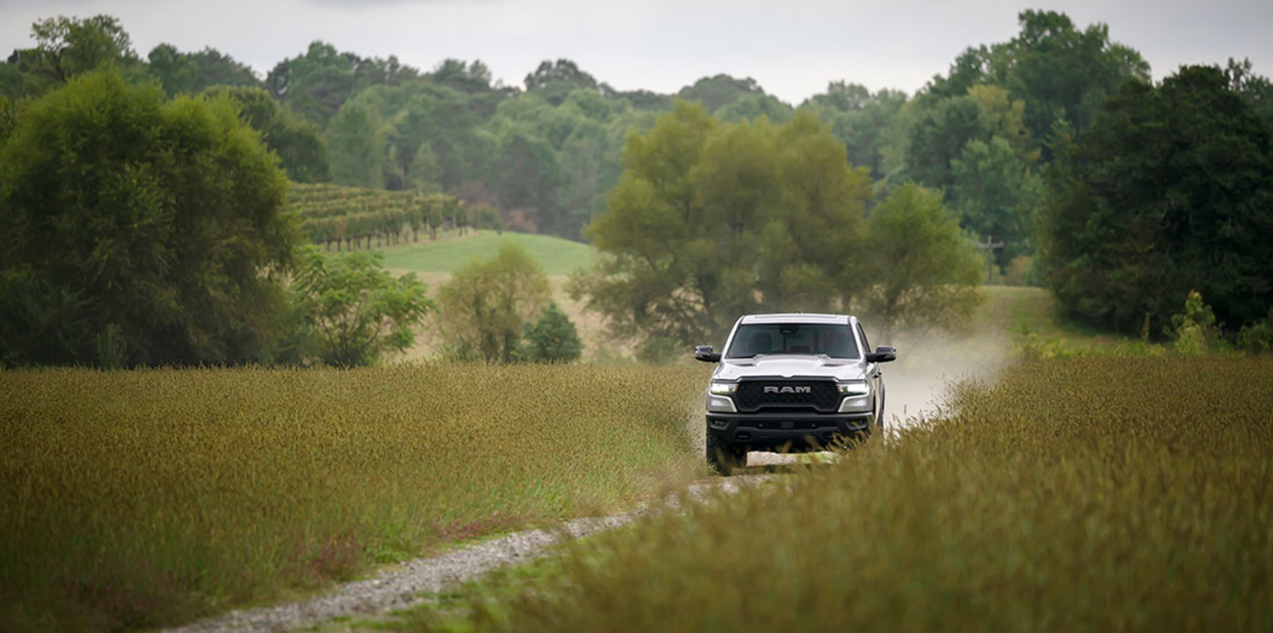 View of a white 2025 Ram 1500 shown driving along an unpaved roadway with trees shown in the distance.