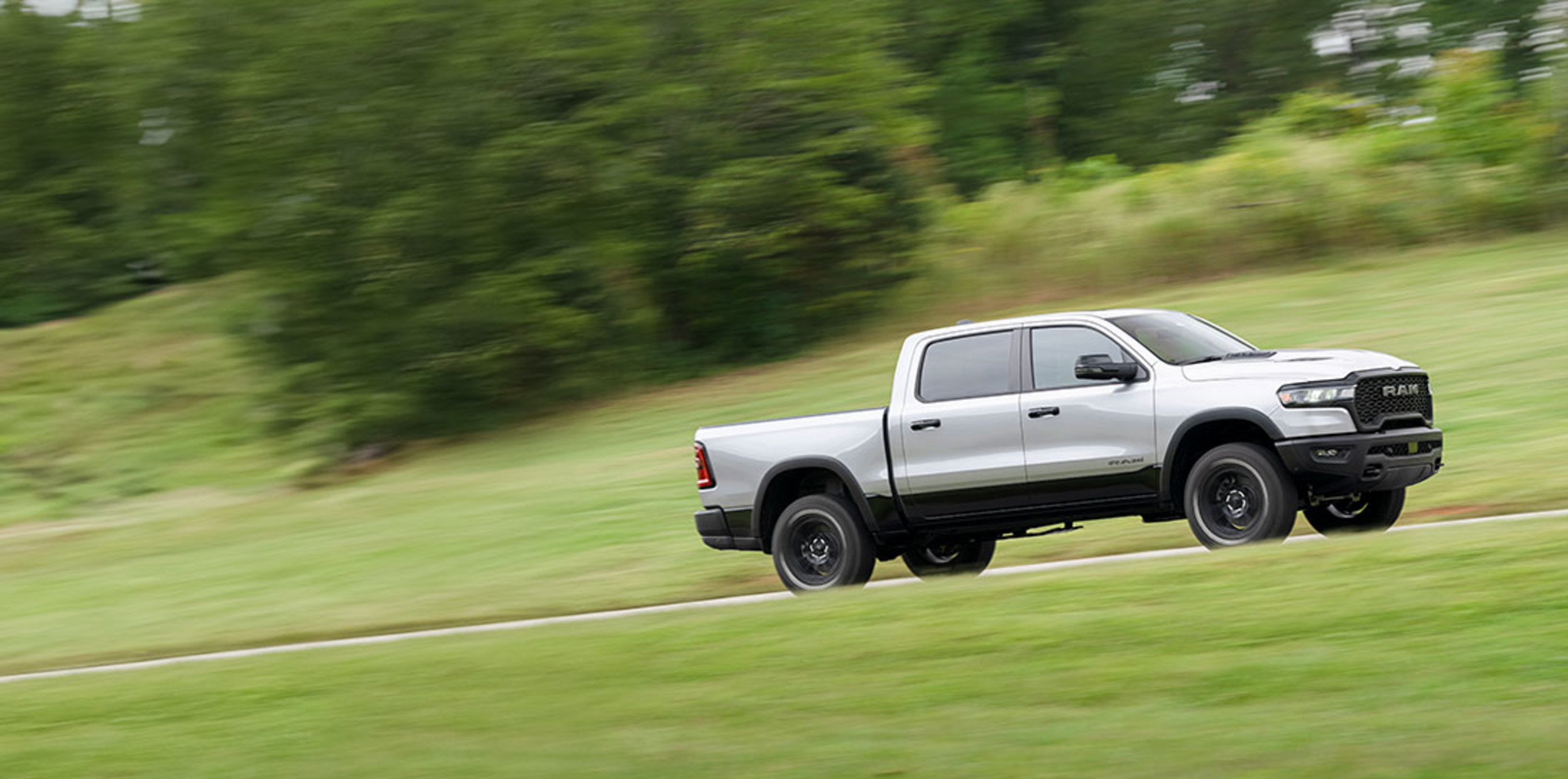 A profile view of a grey 2025 Ram 1500 shown driving on a path with trees shown in the distance.